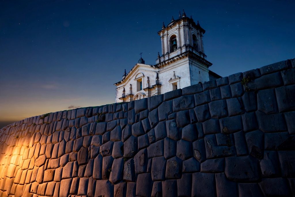 A smooth curved wall of tightly fitted dark Inca stone at dusk, with a pale colonial church rising on top of it.