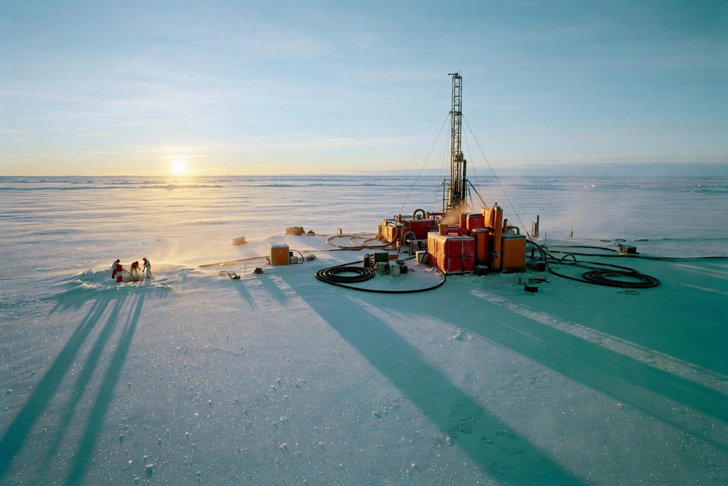 A drilling camp of red containers and a tall mast on the flat Antarctic ice, with researchers in protective suits at the borehole.