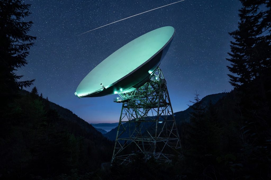 A huge white radio telescope dish pointed at a starry night sky above a dark forest.