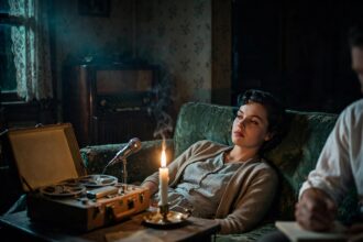 A young woman in 1950s clothing lies on a couch in a dark living room, watching a candle, while a reel-to-reel tape recorder runs beside her.