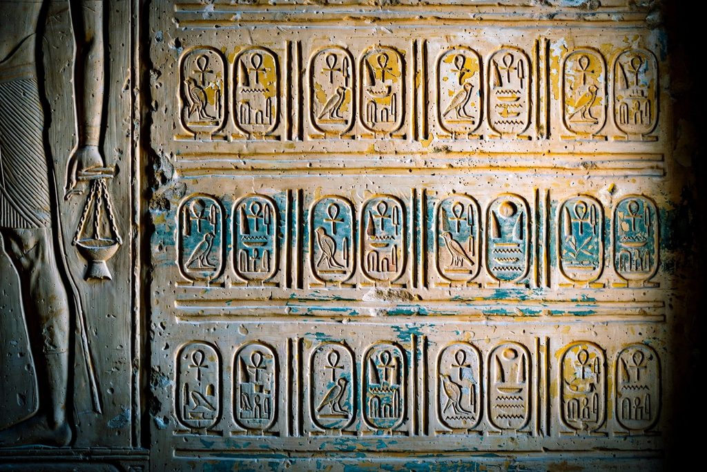Rows of carved royal cartouches on a limestone temple wall, with the feet of a figure holding a censer at the left edge.