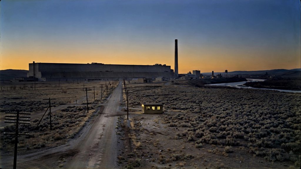 Massive concrete processing buildings and a tall stack on the dry plain beside the Columbia River at dusk.