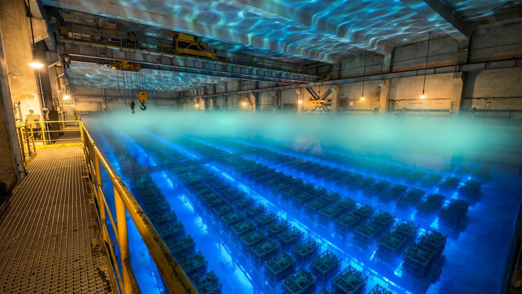 A deep spent fuel pool glowing blue, with fuel racks far below the surface and a crane bridge overhead.