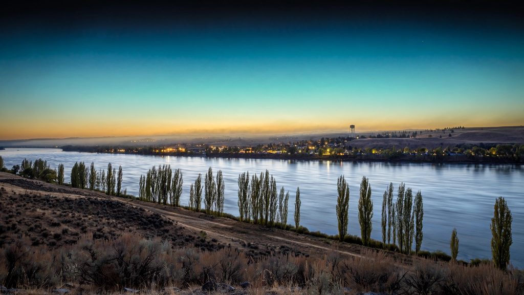 The Columbia River near Richland at dusk, with brown hills, poplars and the lights of the town.